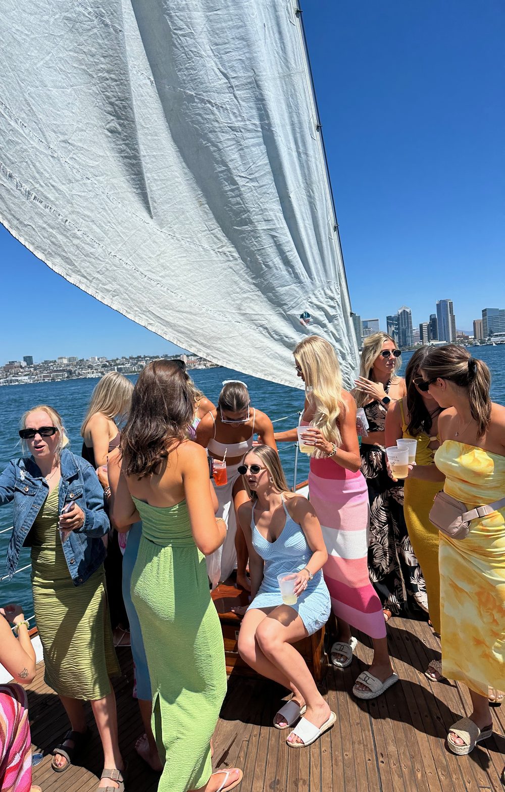 Friends in bright dresses laughing and toasting on JADA's deck under the sail